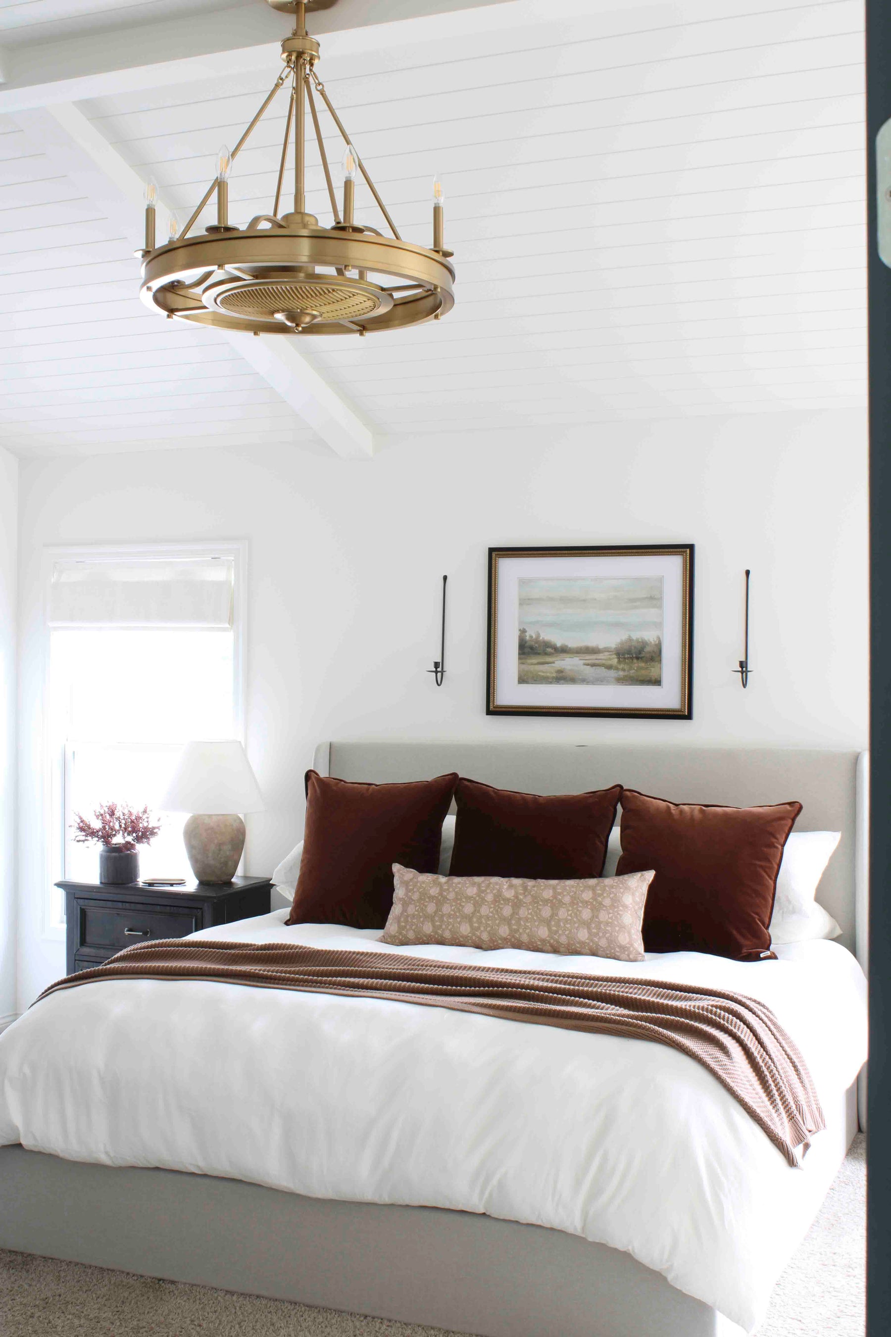 Bright bedroom with a gold fandelier, white shiplap ceiling, gray bed with white bedding, brown pillows, and a brown throw, styled with a black nightstand and landscape artwork above the bed