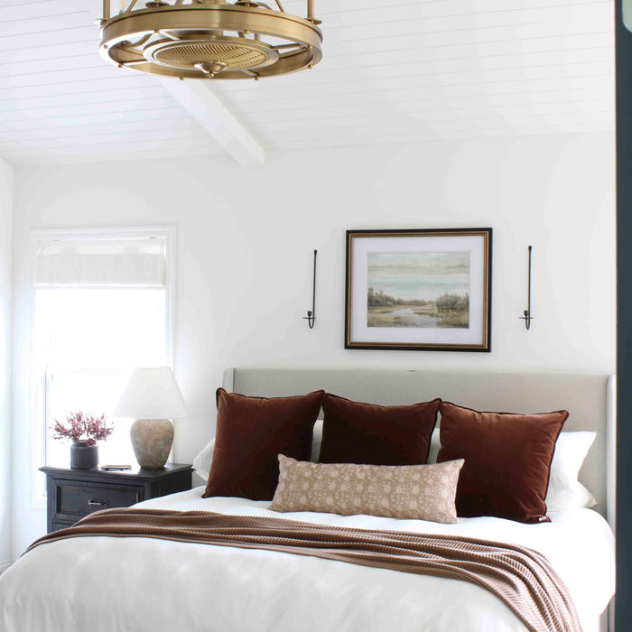Bright bedroom with a gold fandelier, white shiplap ceiling, gray bed with white bedding, brown pillows, and a brown throw, styled with a black nightstand and landscape artwork above the bed