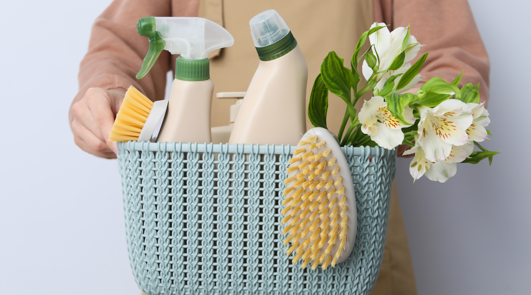 A person holds a light blue basket filled with cleaning supplies and flowers
