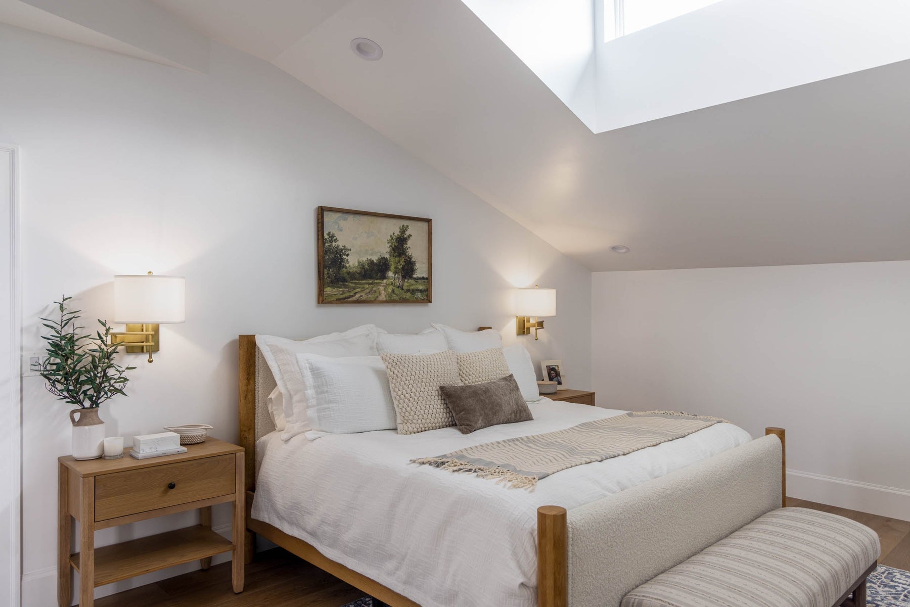 Attic-style bedroom with sloped ceilings, wall-mounted sconces, neutral bedding, and wood furniture.
