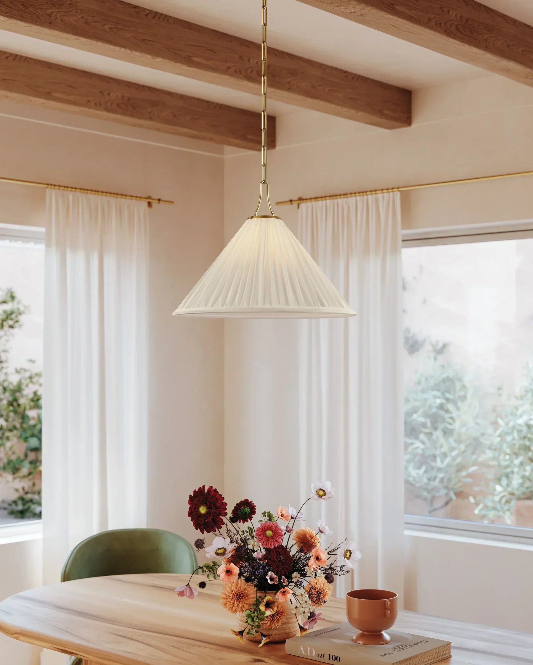 A pleated white shade pendant hangs over a dining room table in front of two large windows