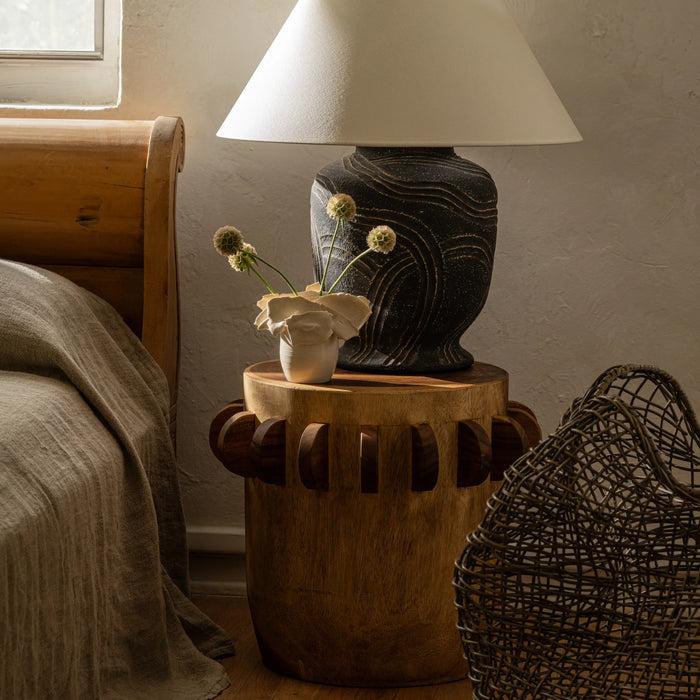 Large cement table lamp sits on a wood nightstand next to a warm wood bed