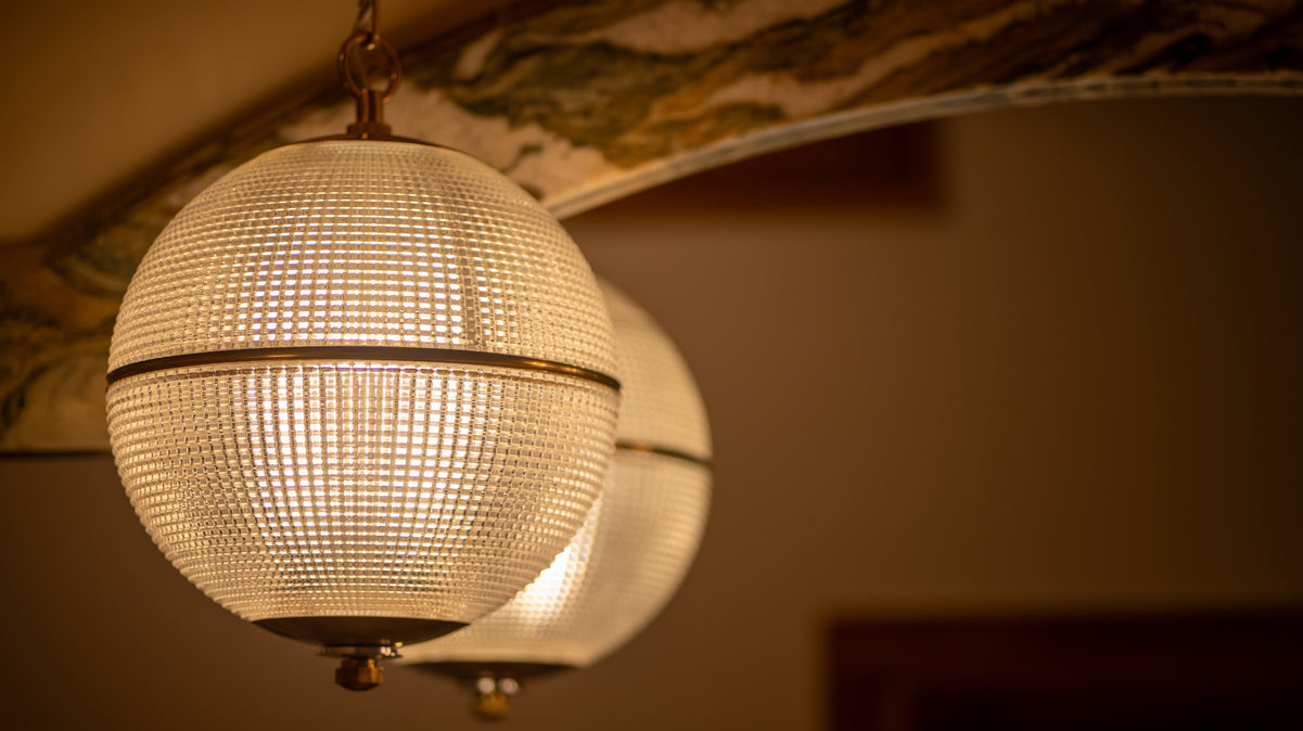 Two spherical pendant lights with textured glass shades and warm brass accents hang in front of a marble backsplash, casting a soft, warm glow that creates an elegant and inviting atmosphere.