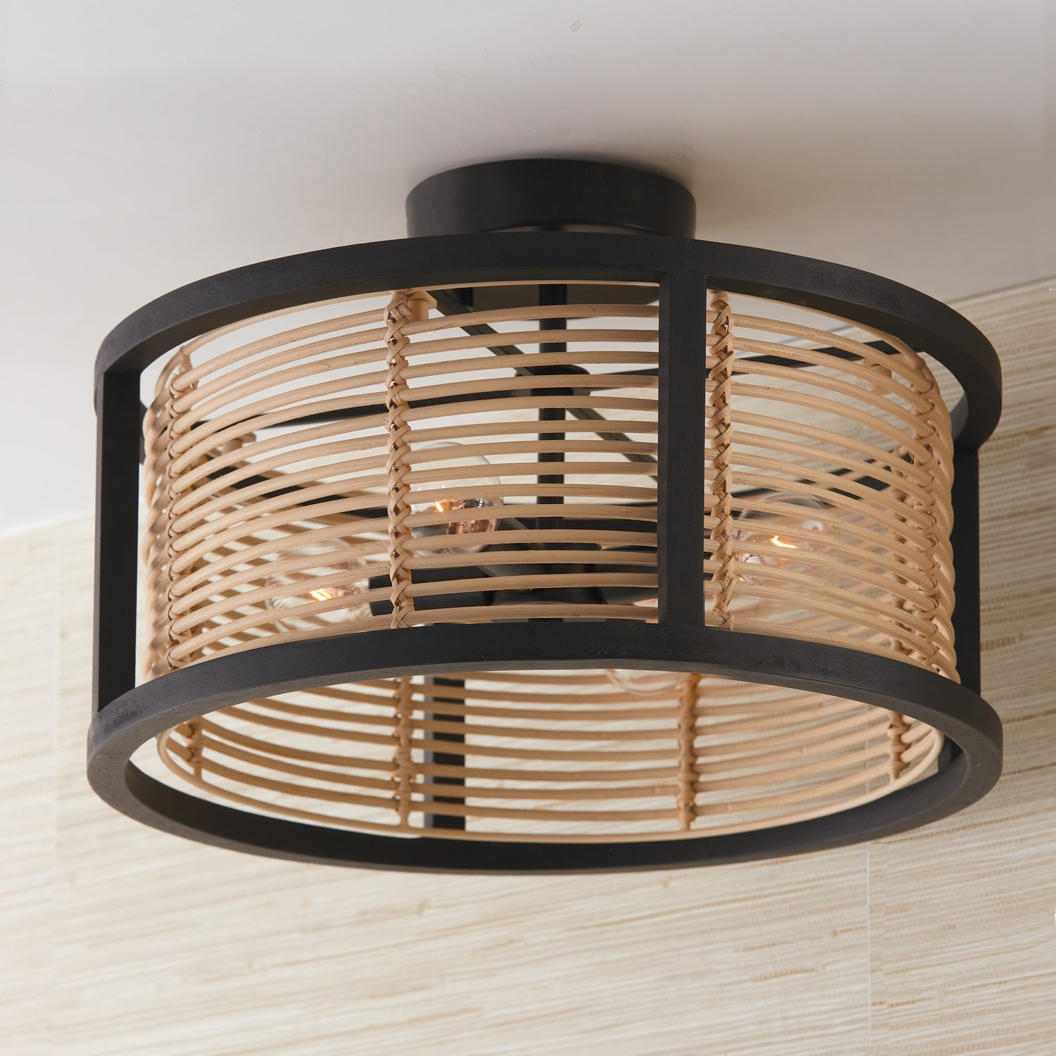 Round semi-flush ceiling light fixture with a black metal frame and woven light wood/rattan drum shade encasing visible Edison-style bulbs.