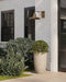Decorative outdoor planters with greenery in front of a building with a wall-mounted light fixture.