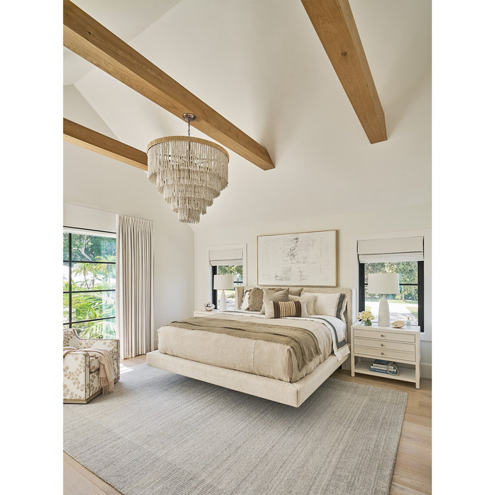 Bright and cozy neutral bedroom featuring vaulted ceilings with exposed wood beams, layered cream bedding, a statement chandelier, large windows with soft drapery, light wood flooring, and a textured area rug, creating a warm and luxurious modern farmhouse retreat.