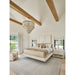 Bright and cozy neutral bedroom featuring vaulted ceilings with exposed wood beams, layered cream bedding, a statement chandelier, large windows with soft drapery, light wood flooring, and a textured area rug, creating a warm and luxurious modern farmhouse retreat.
