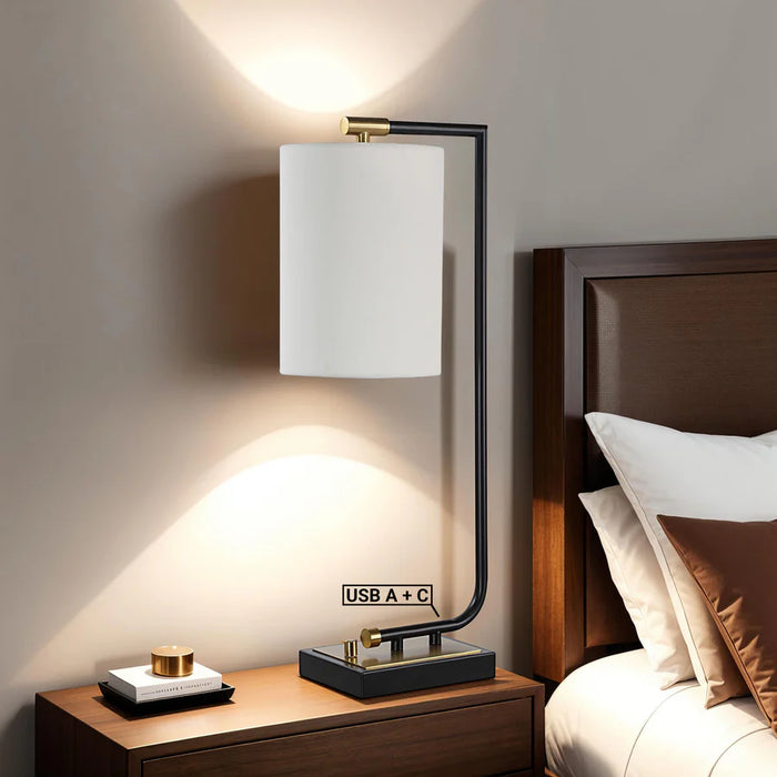 Modern black and gold bedside table lamp with a white cylindrical shade, illuminated beside a wooden bed and nightstand.
