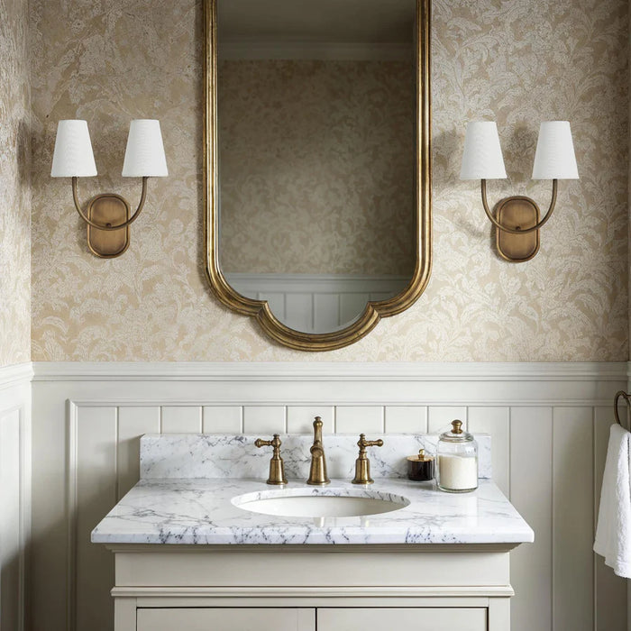 Classic bathroom vanity with marble countertop, gold-framed mirror, and matching brass double wall sconces with white shades.