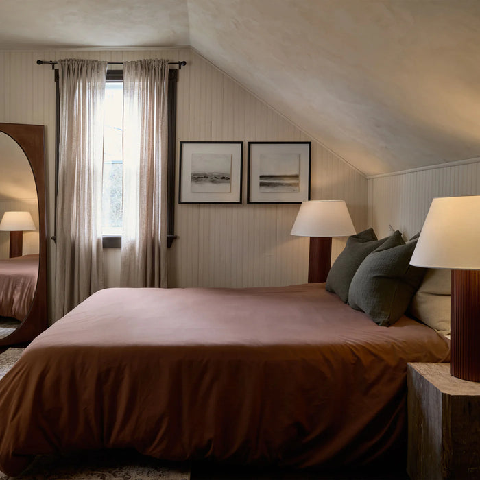 California modern cozy bedroom featuring warm terracotta bedding, soft neutral walls with vertical paneling, layered pillows, ambient table lamps, sheer curtains, framed artwork, and natural textures creating a warm and inviting bedroom retreat.