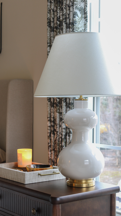 White lamp on a wooden surface with a candle and decorative tray in the background.
