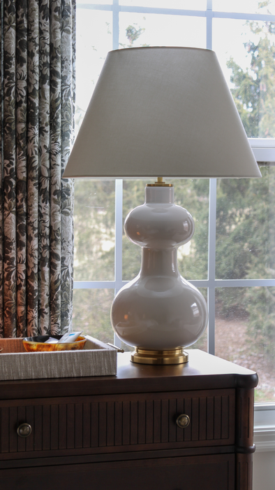 White lamp with a beige shade on a wooden dresser by a window