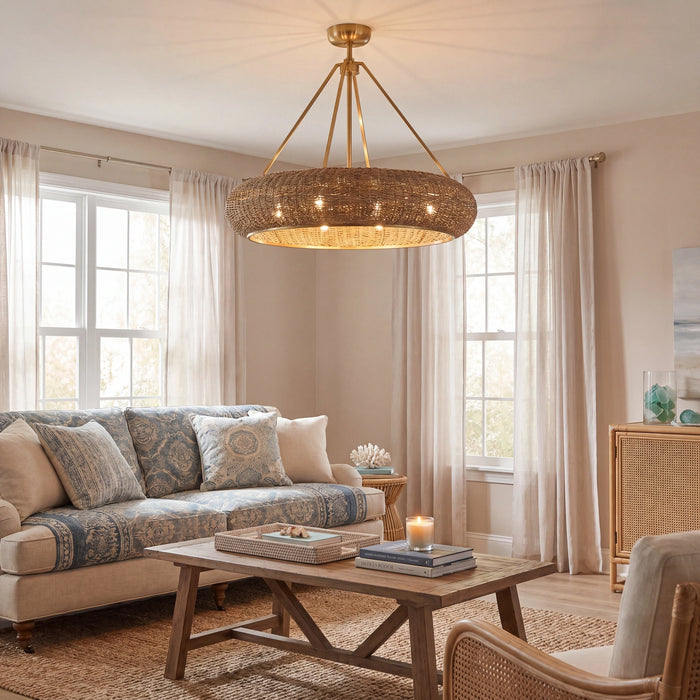 A large circular woven pendant chandelier with a brass frame hangs in a bright, modern living room with beige walls, sheer curtains, a patterned sofa, and a wooden coffee table.