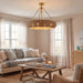 A large circular woven pendant chandelier with a brass frame hangs in a bright, modern living room with beige walls, sheer curtains, a patterned sofa, and a wooden coffee table.