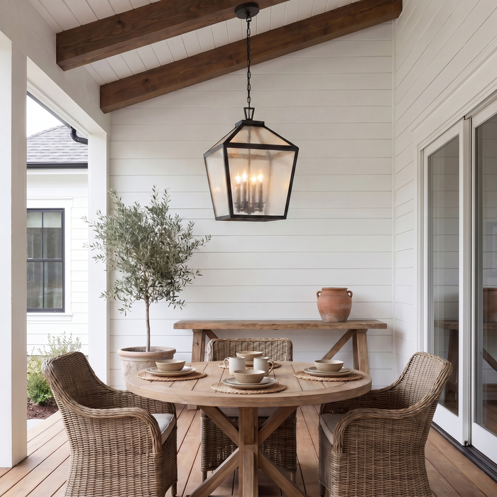 A black metal hexagonal outdoor pendant light with glass panels hangs over a round wooden dining table on a porch with wicker chairs, white shiplap walls, and a potted tree.
