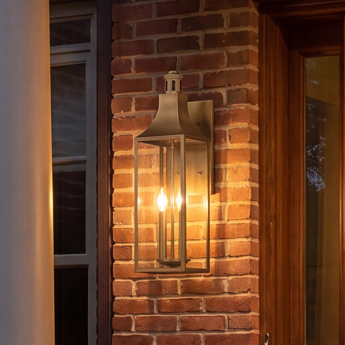 A warm-toned outdoor wall sconce with a brass-finished metal frame and glass panels, mounted on a red brick pillar next to a wooden door.