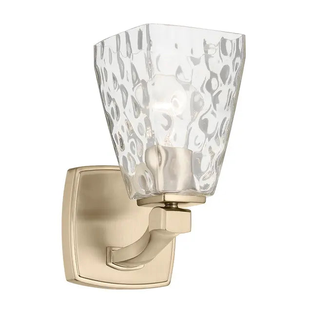 A wall-mounted light fixture with a square brushed brass or gold-toned metal backplate and curved arm, paired with a clear textured glass shade. The shade features a hammered or wavy pattern that diffuses the light.