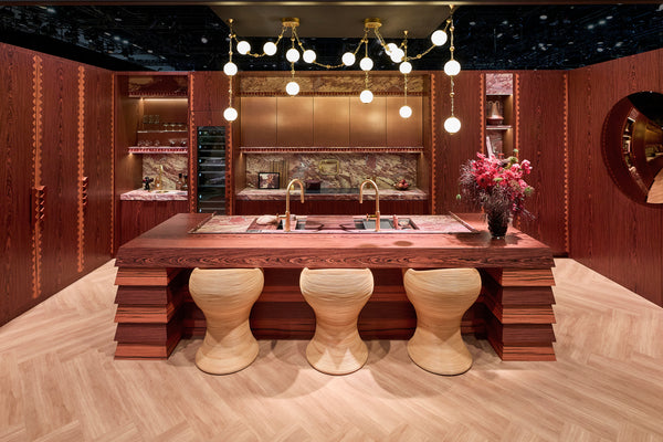 Modern kitchen with wooden island and stools in a stylish interior setting. Large swagged, modern chandelier in brass hangs from ceiling.