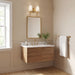 A modern floating bathroom vanity with a wood cabinet, white countertop, and gold faucet, shown in a styled beige bathroom with a gold-framed mirror, wall sconce, towel bar, and window.