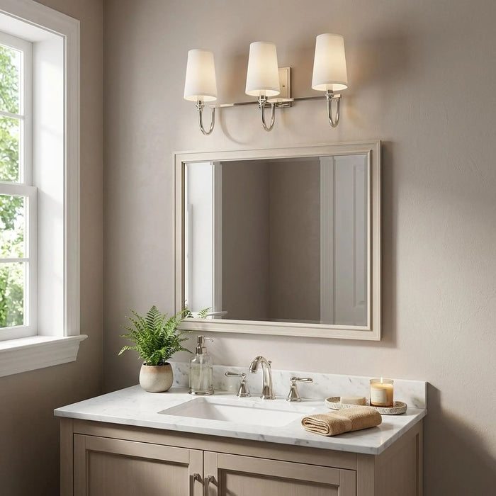 A three-light wall sconce with cream fabric shades and a brushed nickel frame is mounted above a rectangular wall mirror in a bathroom setting with a marble countertop and beige cabinetry.