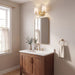 A modern bathroom vanity setup with a medium brown wooden cabinet, white marble countertop, white sink, a gold faucet, a matching wall mirror, and a gold sconce light fixture above. The room has beige walls, a window with sheer curtains on the left, and a gold towel ring with a white towel on the right.