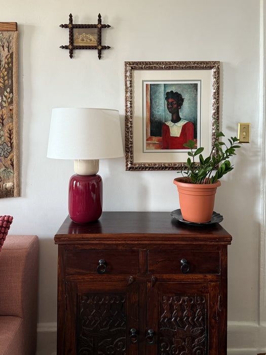 Vintage-style table lamp with a white shade on a carved wood cabinet, styled with framed artwork, a small potted plant, and eclectic wall decor against a neutral wall.
