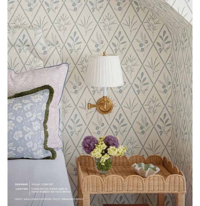 A styled bedroom corner featuring a cream wallpaper with a muted blue botanical diamond pattern, a white pleated wall lamp with a brass mount, a wicker side table with flowers, and part of a bed with patterned pillows.