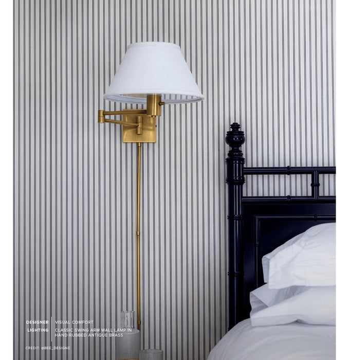A brass wall lamp with a white conical shade mounted on a striped wall beside a bed with white bedding and a dark wooden headboard.