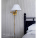 A brass wall lamp with a white conical shade mounted on a striped wall beside a bed with white bedding and a dark wooden headboard.