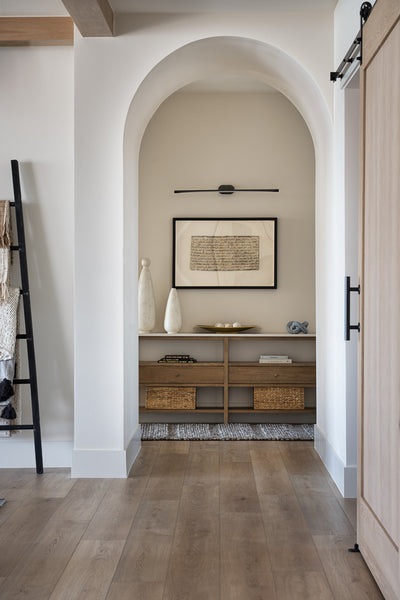 Arched hallway nook with a built-in wood console, decorative vases and artwork, wood flooring, and a minimalist picture light above the shelf.