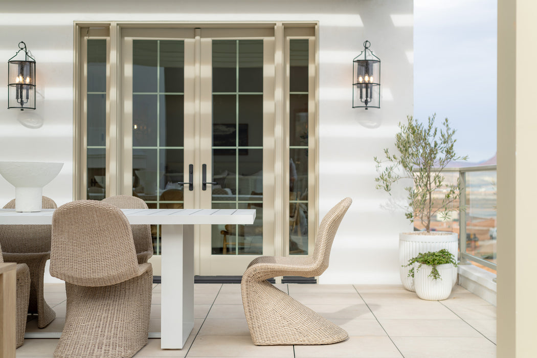 Outdoor patio dining area with woven chairs around a white table, French doors, potted greenery, and black lantern-style wall sconces.