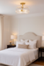 A cozy, neutral-toned bedroom with a light beige upholstered headboard, crisp white bedding, and matching bedside tables topped with brass lamps. A brass and ribbed-glass semi-flush mount light fixture is centered on the ceiling, casting a warm, soft glow over the room.