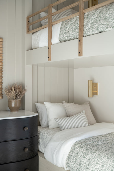 Bunk bedroom with a lower bed dressed in neutral bedding, an upper wood bunk rail, vertical shiplap walls, and a brass wall sconce for reading light.