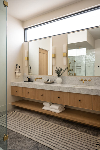 Modern bathroom with a long wood vanity, marble countertop, double mirrors, brass wall sconces, and warm accent lighting above dual sinks.