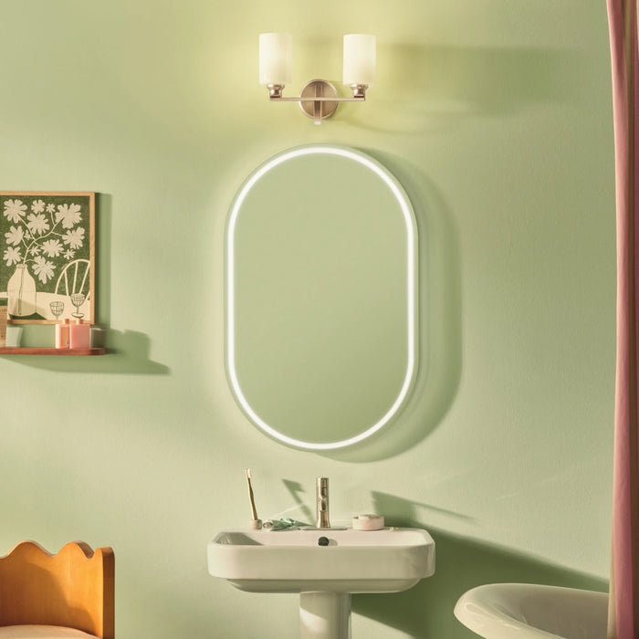 An oval LED-backlit bathroom mirror mounted on a sage green wall above a white pedestal sink, with a wall-mounted two-light sconce above it.