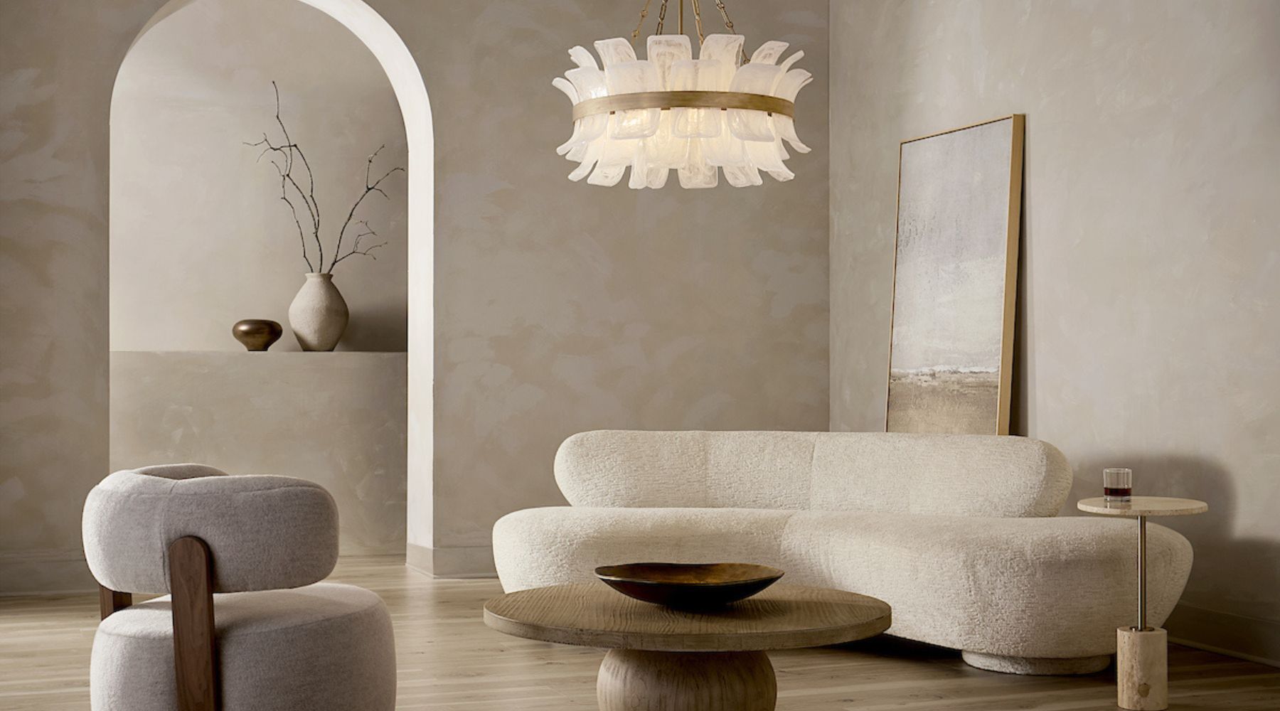 Neutral-toned modern living room with a sculptural cream sofa, rounded accent chair, wood coffee table, arched wall niche, and an elegant layered glass chandelier overhead.