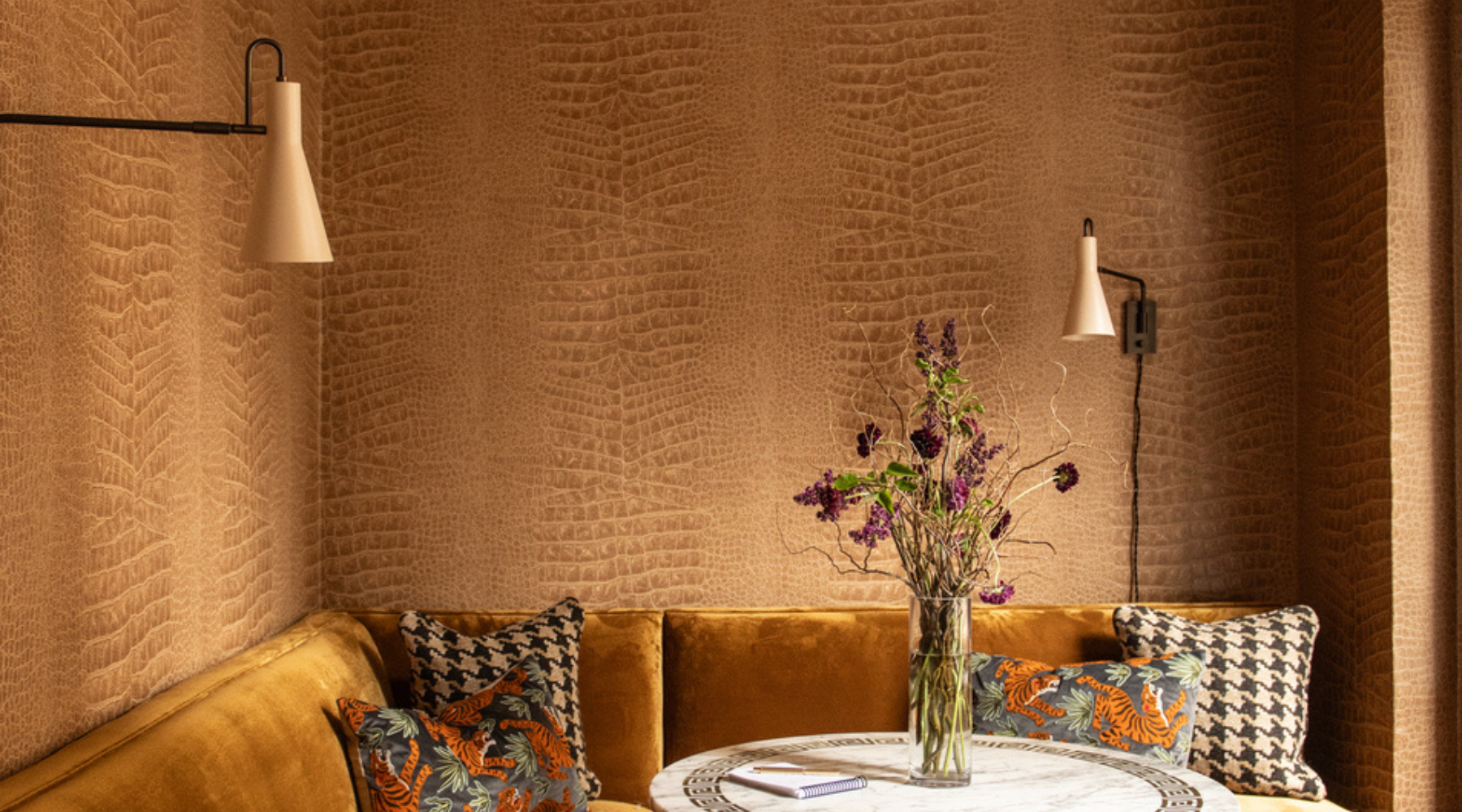 A cozy interior corner with a mustard velvet sofa, patterned pillows, a round marble-top table with a vase of dried purple flowers, and textured warm brown walls with two wall sconces.