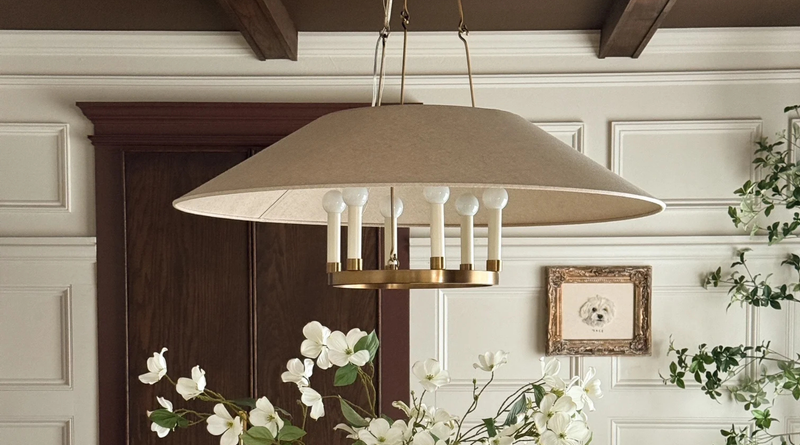Close up photo of a six light modern brass chandelier with a tan linen shade. Hangs from a brown ceiling over a dining table with a white floral arrangment sitting on top