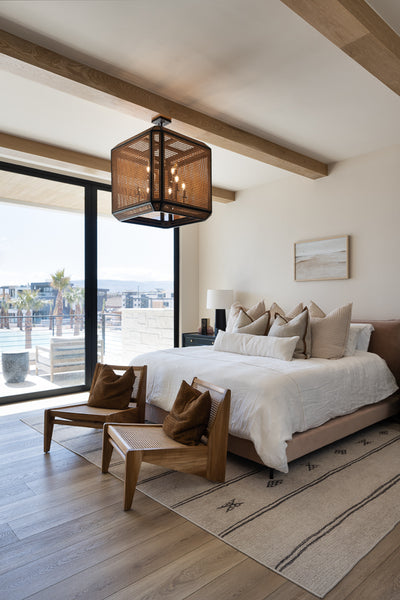 Modern bedroom with a woven square pendant light hanging above a low-profile bed, exposed wood ceiling beams, neutral bedding, and large sliding glass doors.