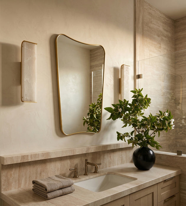 A modern bathroom vanity scene with a large asymmetrical gold-framed mirror, matching wall sconces, a stone countertop, folded towels, and a black vase holding a leafy green plant.