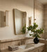 A modern bathroom vanity scene with a large asymmetrical gold-framed mirror, matching wall sconces, a stone countertop, folded towels, and a black vase holding a leafy green plant.