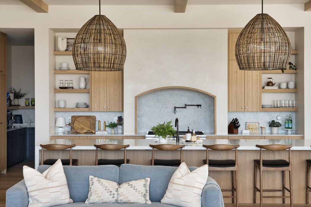 Open-concept kitchen with two large woven pendant lights hanging over a marble island, light wood cabinetry, open shelving, and a soft blue sofa in the foreground.