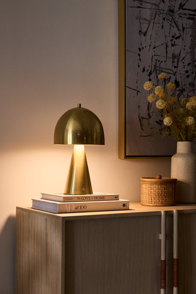 Gold table lamp on a wooden surface with books and a vase in the background
