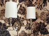 Wall sconces with white lampshades on a floral wallpapered wall.