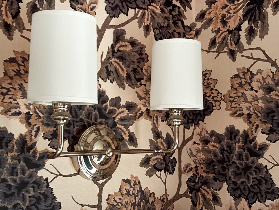 Wall sconces with white lampshades on a floral wallpapered wall.