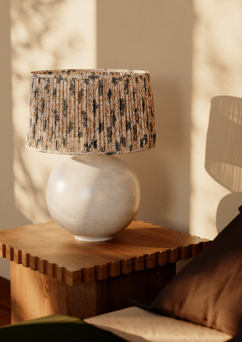 A table lamp with a textured beige and black patterned drum shade and a round off-white ceramic base, styled on a wooden side table in a warm, sunlit room.