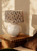 A table lamp with a textured beige and black patterned drum shade and a round off-white ceramic base, styled on a wooden side table in a warm, sunlit room.