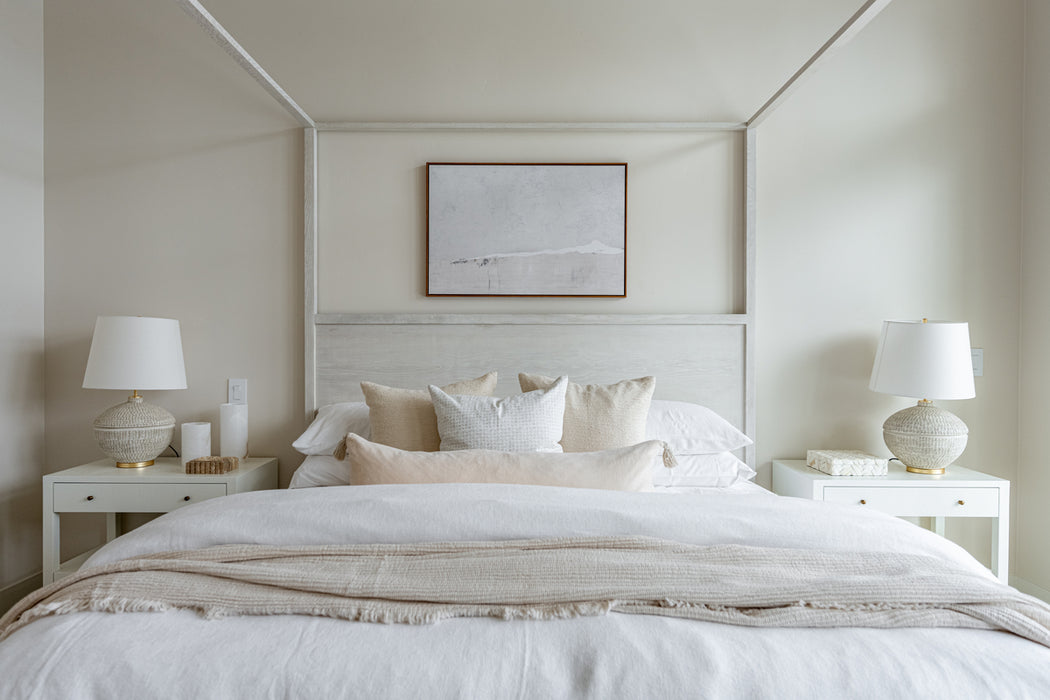 Serene bedroom with a white upholstered bed, layered neutral bedding, matching nightstands with table lamps, and soft artwork above the headboard.