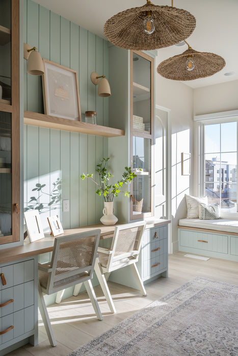 Sunlit home office nook with sage green paneling, built-in desk and shelving, woven pendant lights, wall sconces, and a cozy window seat.