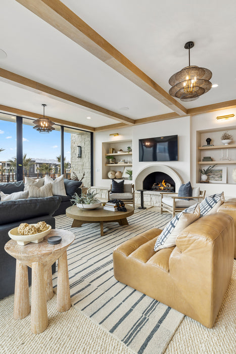 Bright living room with woven pendant lights, exposed wood ceiling beams, an arched stone fireplace, built-in shelves, and layered neutral seating with leather and fabric sofas.